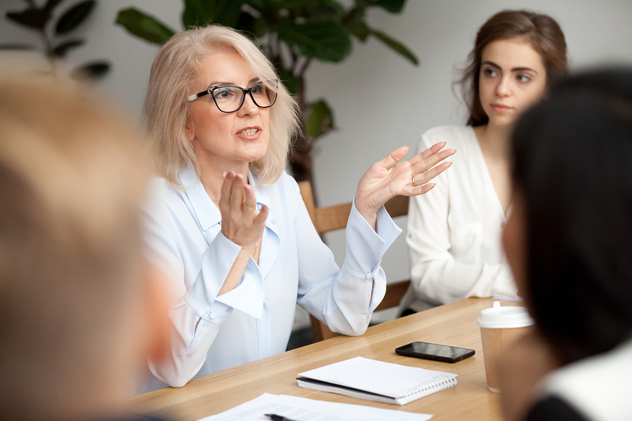 Attractive aged businesswoman, teacher or mentor coach speaking to young people, senior woman in glasses teaching audience at training seminar, female business leader speaker talking at meeting